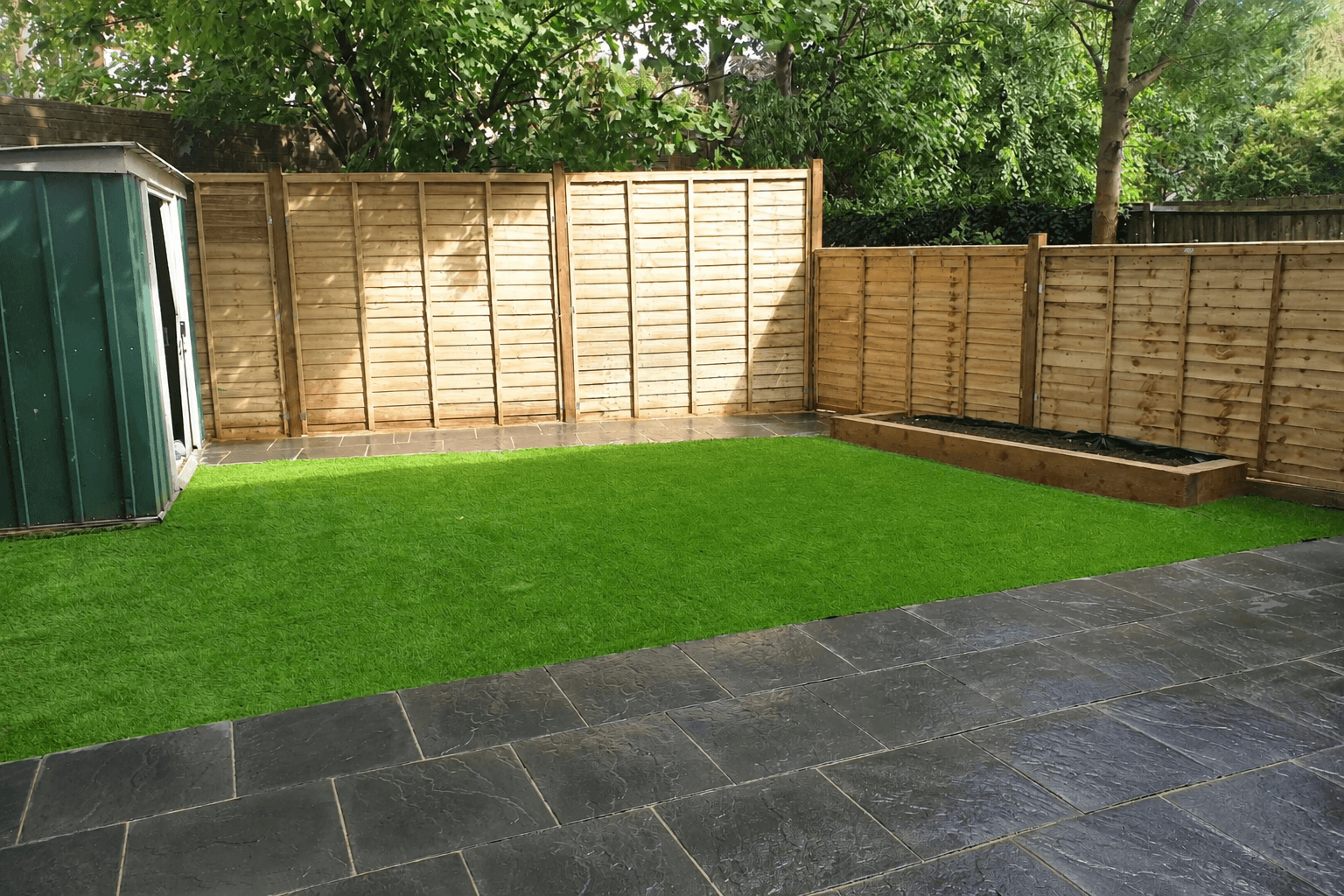 Landscaped back garden in South East London featuring a vibrant artificial grass lawn, a large dark slate patio in the foreground, new wooden panel fencing, a green metal garden shed on the left, a raised timber planter bed along the right fence, and mature trees visible above the fence line