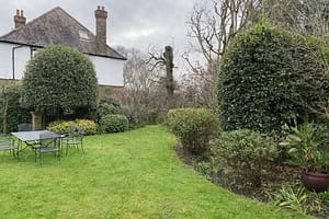 Neat backyard garden with green lawn, patio table and chairs, and mature shrubs beside a white house on a cloudy day